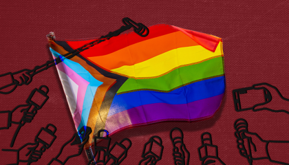 A progress pride flag in front of a dark red background surrounded by press microphones
